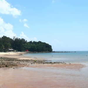 A quiet tropical beach in Sarawak at low tide, with calm waters, a sandy shoreline, luch green hills, and clear blue skies.